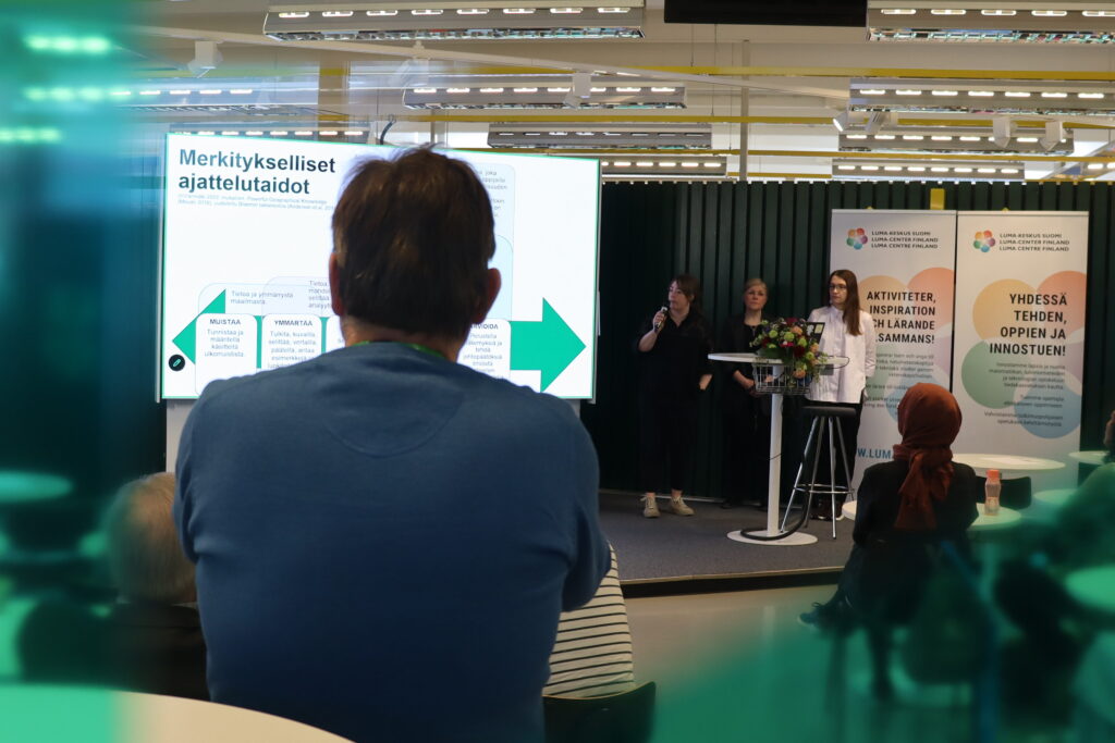 Kuvassa vihreäsävyitteisen ikkunan takaa näkyviä ihmisiä sekä lava, jolla seisoo 3 henkilöä. Yksi heistä puhuu mikrofoniin. Vierellä iso näyttö, jossa näkyy kaavio ja sen yllä otsikko Merkitykselliset ajattelutaidot.