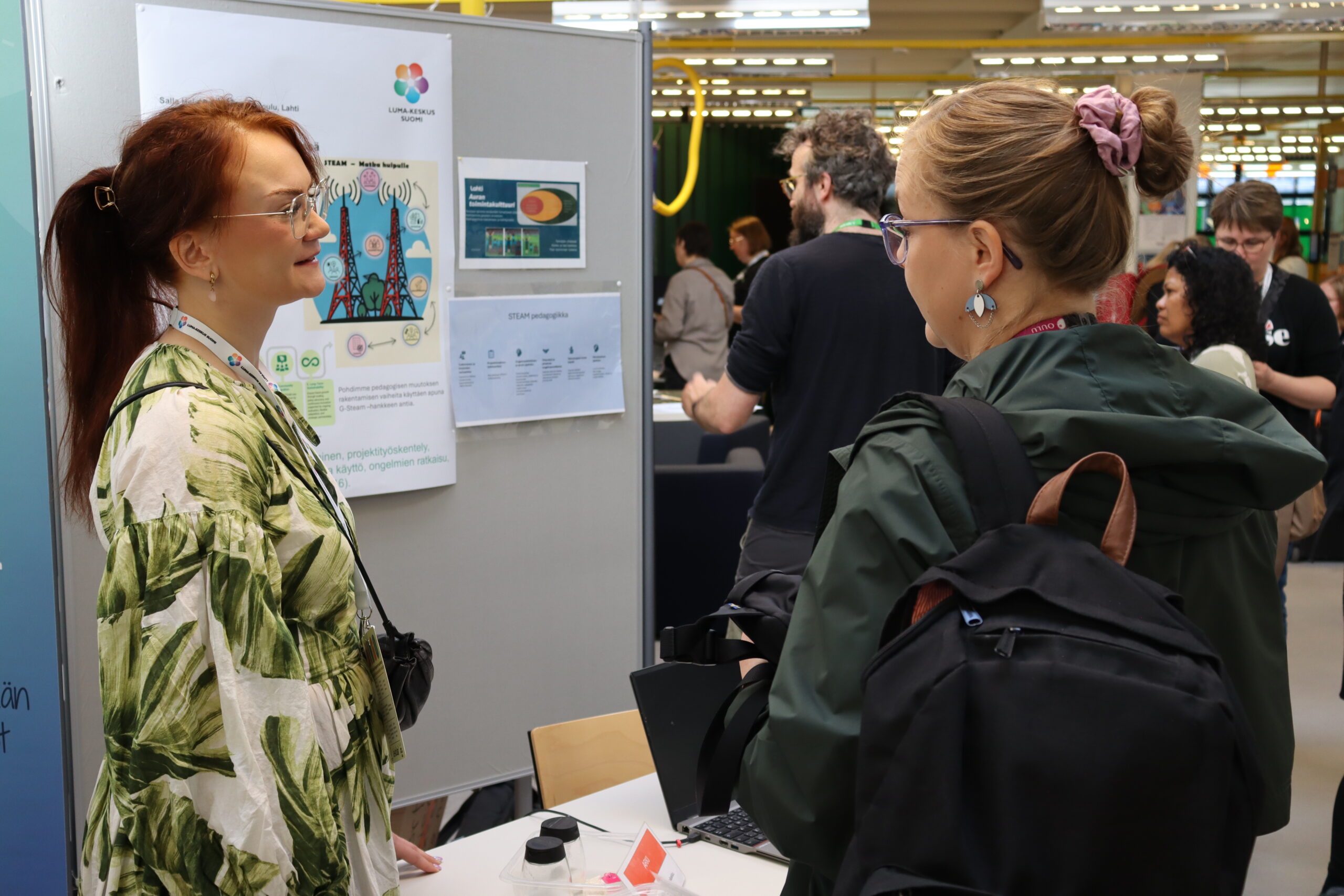 Kuvassa näkyy messupiste, jolla on posteri STEAM-pedagogiikasta. Messupisteen edustaja keskustelee osallistujan kanssa. Taustalla näkyy muista pisteitä, joilla käydään vastaavia keskusteluja.