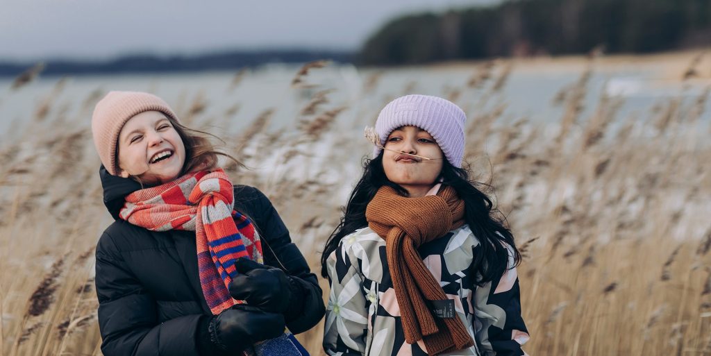 Kuvituskuvassa valokuva kahdesta 8-vuotiaasta tytöstä ulkovaatteissa syksyisellä rannalla. Toinen on tehnyt järviruokosta viikset ja toinen nauraa.