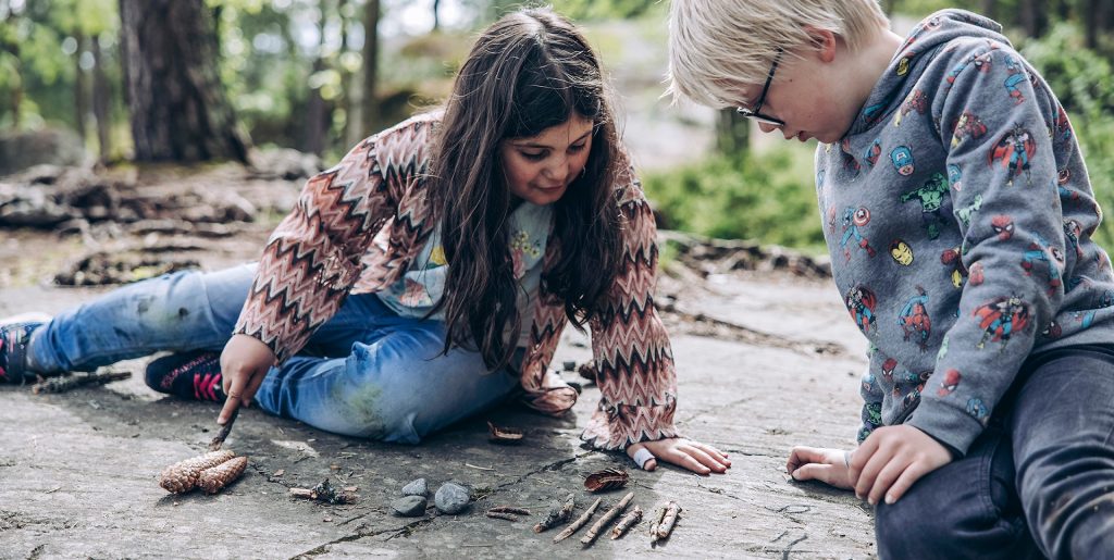 Kuvassa työttä ja poika ratkovat matemaattista tehtävää kävyillä ja kivillä kalliolla istuen.