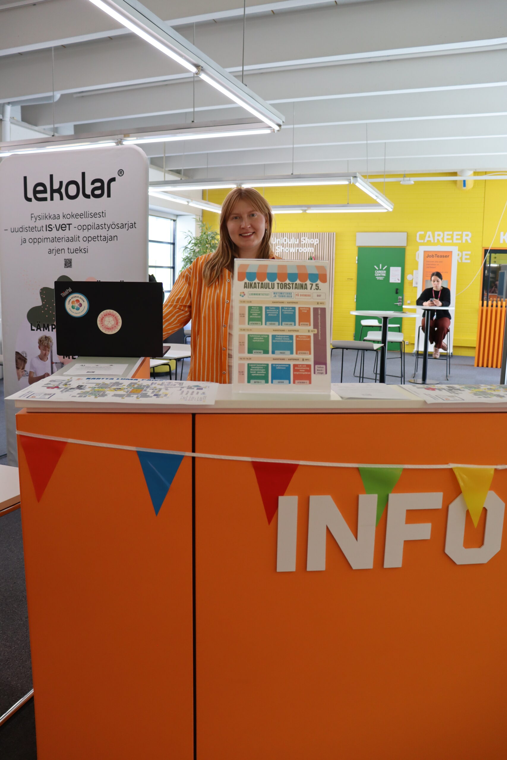 Kuvassa INFO-tekstillä varustetulla asiakaspalvelupisteellä seisova nainen, Oona Kiviluoto verkoston hallinnosta. Taustalla rollup, jossa lukee lekolar. 